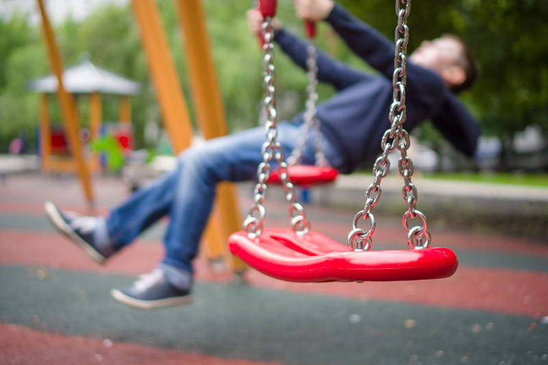 A close up of a red swing with a plastic seat and metal chains with a child in soft focus in the background.